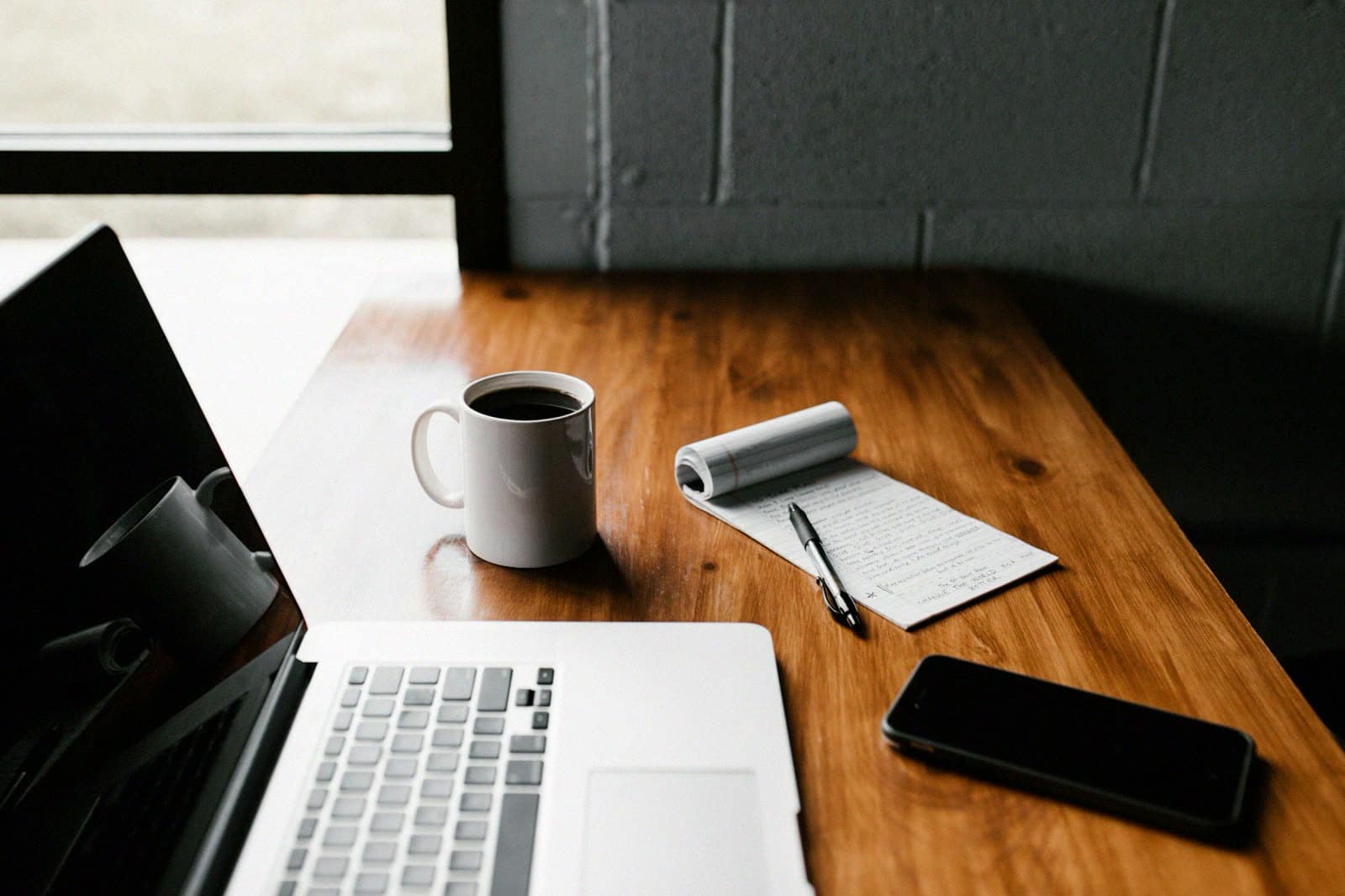 A laptop, notepad, phone and mug of coffee on a wooden desk