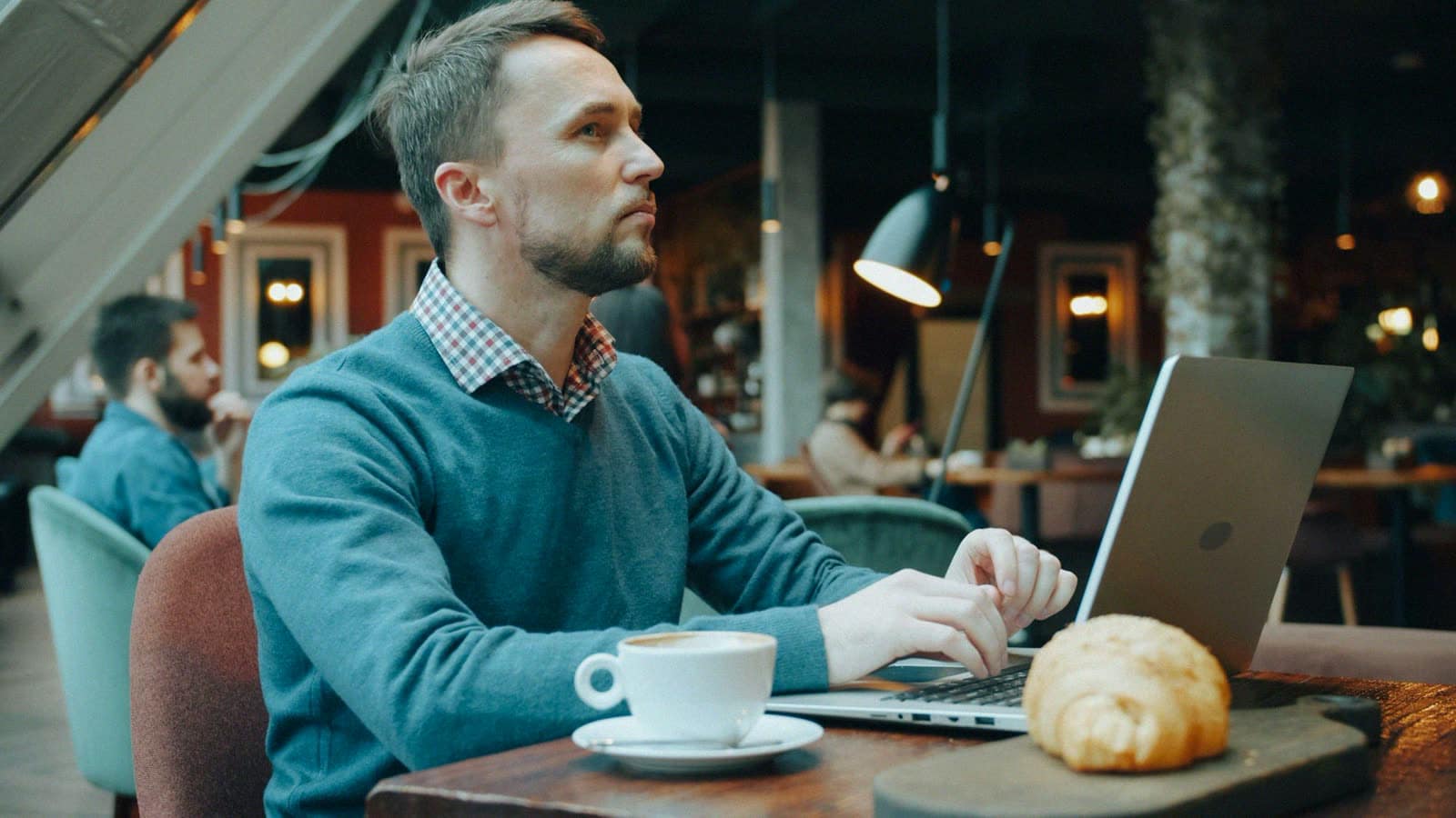 A man working on a laptop in a cafe beside a coffee and a croissant
