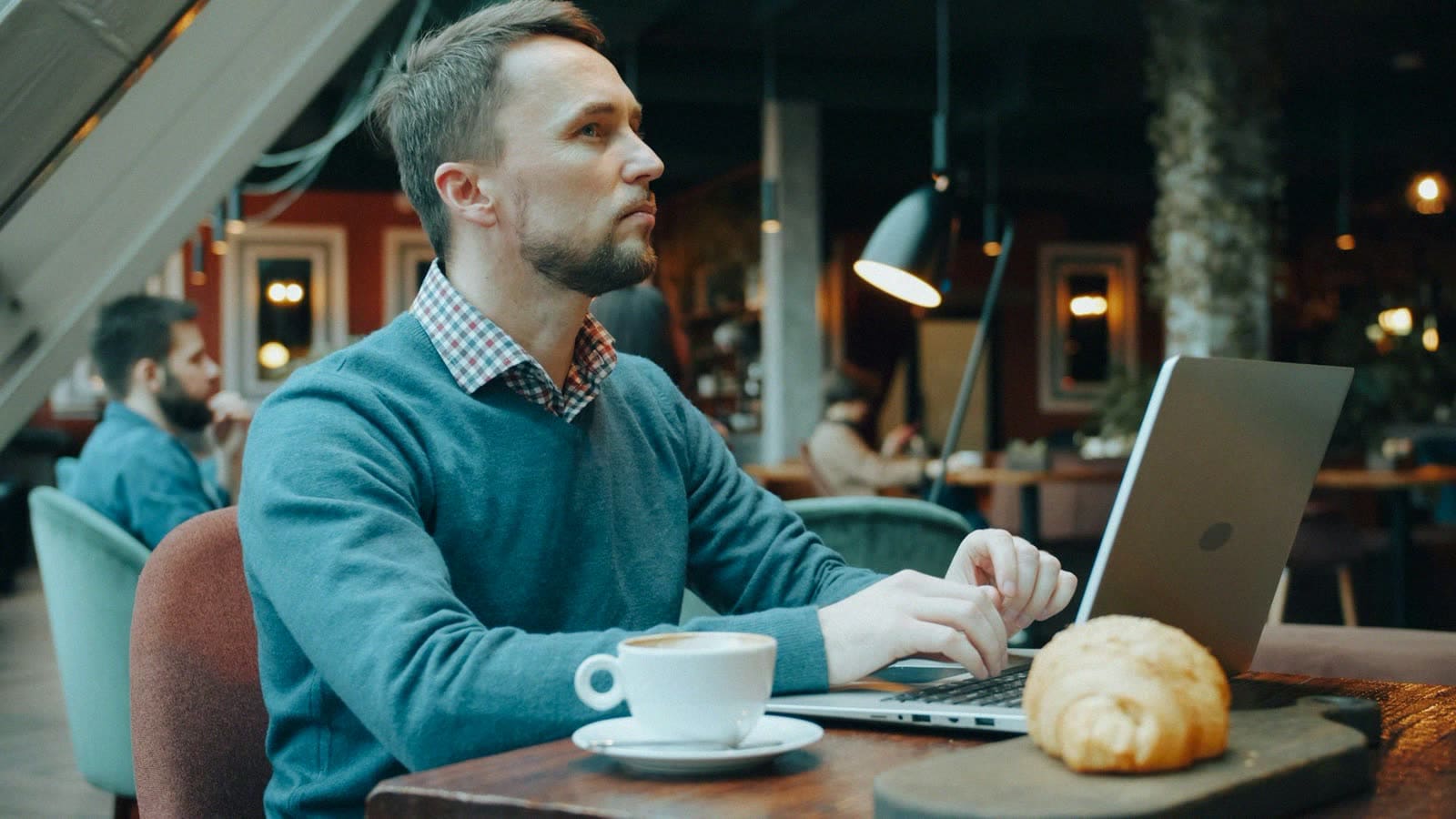 A man working on a laptop in a cafe beside a coffee and a croissant